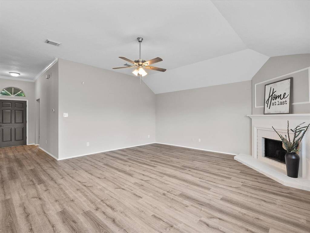 Spacious living room with light LVT flooring, neutral walls, and vaulted ceiling. Features a ceiling fan, cozy fireplace, and a welcoming entryway with a dark door.