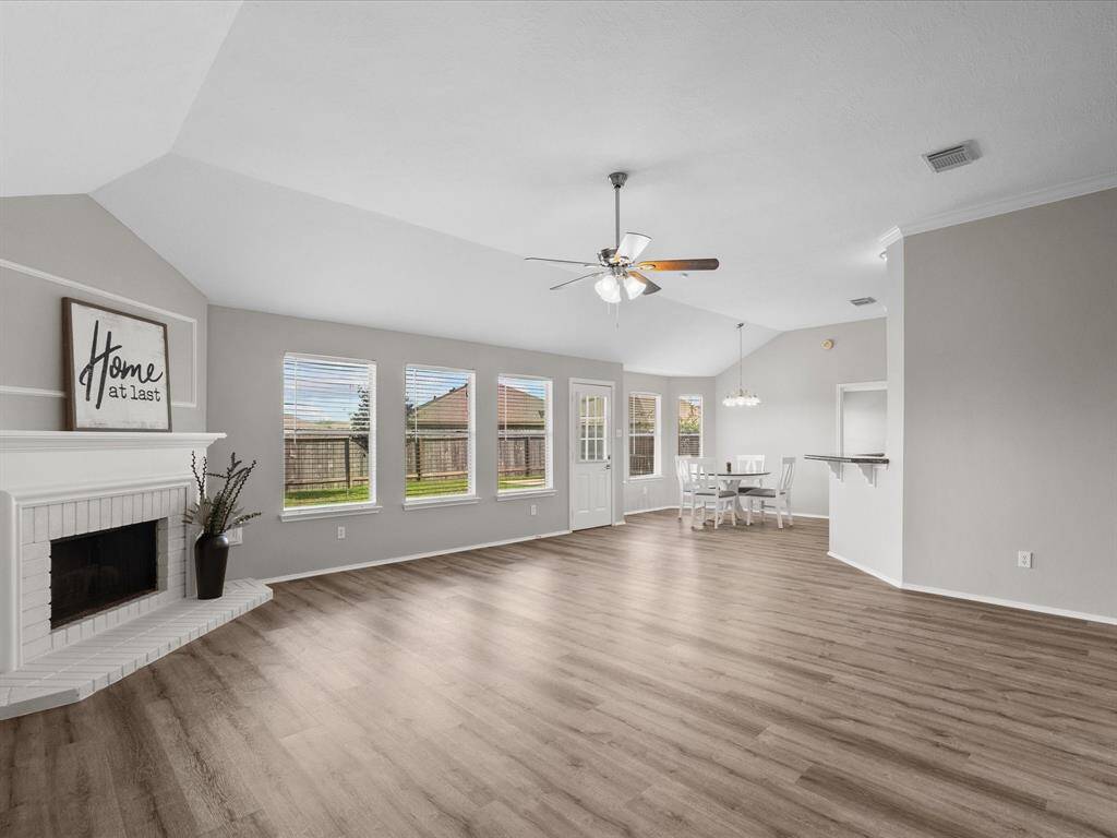This spacious living area features a vaulted high ceiling and plenty of natural light from multiple windows. It includes a cozy white brick fireplace and an open layout that connects to a breakfast space, making it ideal for entertaining.