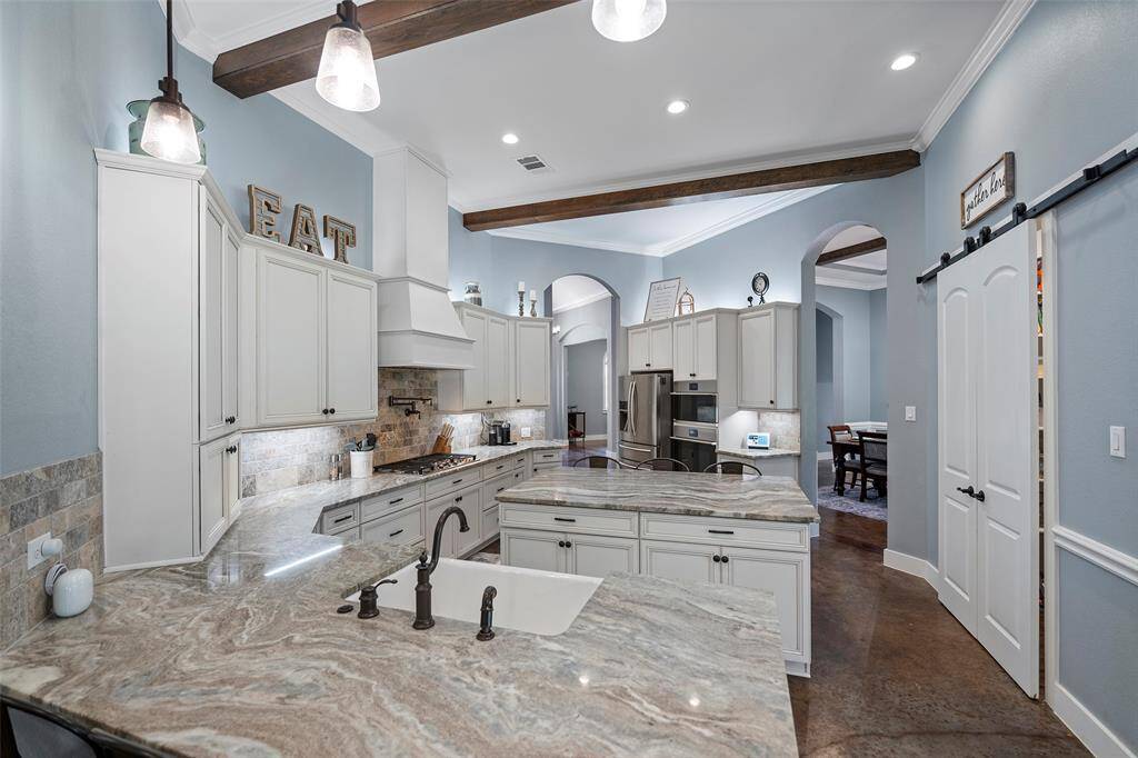 This open layout features a chef’s island kitchen w/custom cabinets, a pot filler, under/above cabinet lighting, w/barn door walk-in pantry with views of the spacious family room w/wood burning fireplace wooden beams, & large picture windows with beautiful views to the back acreage.