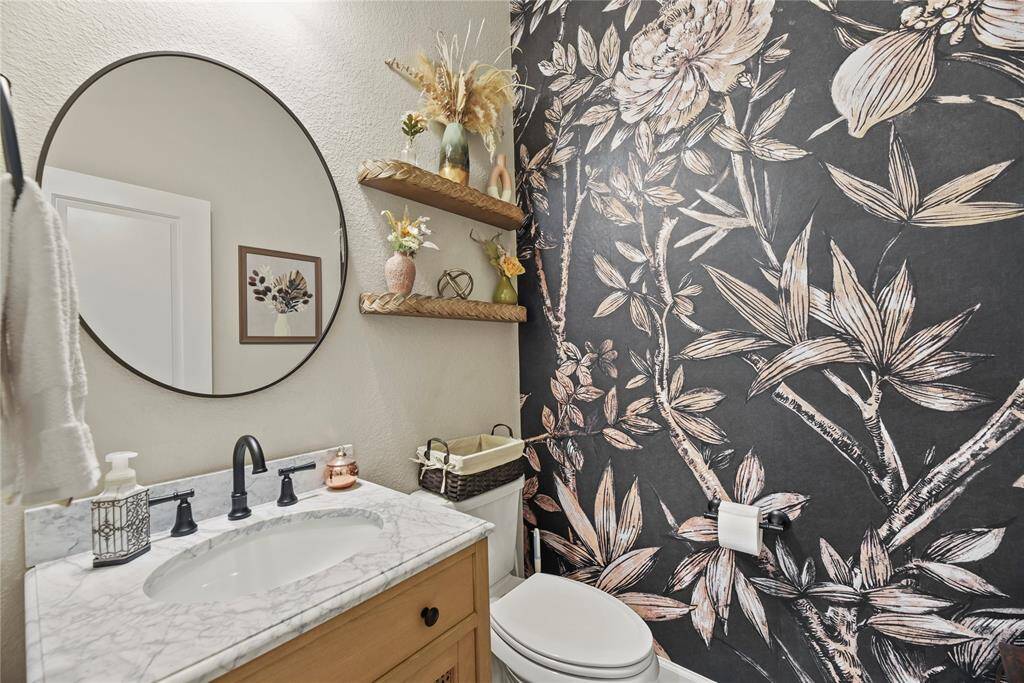 This half bathroom features a stylish floral accent wallpapered wall, a framed mirror above a marble-topped vanity, and decorative shelves. The overall design is modern and perfect for this chic home.