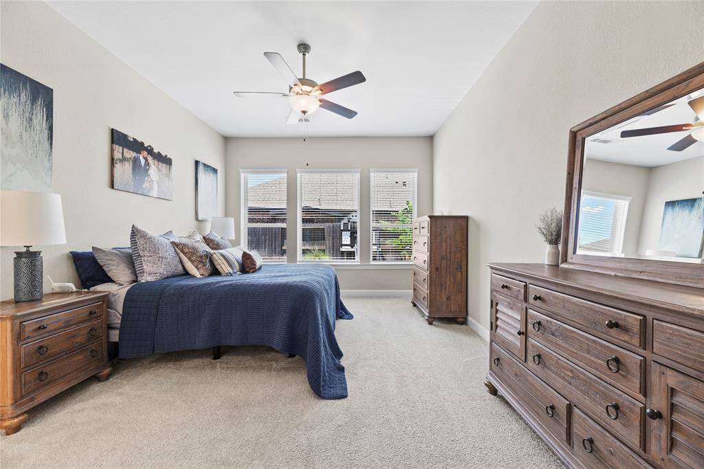 This spacious bedroom features neutral walls and carpet, with ample natural light from three large windows and includes a ceiling fan.