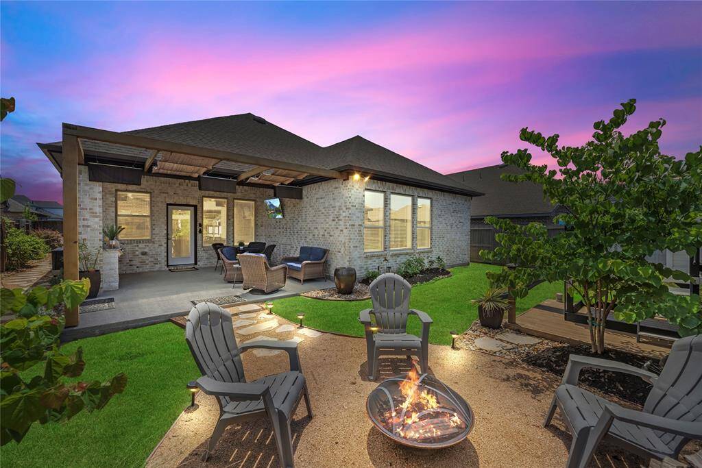 This photo showcases a cozy backyard with a covered patio, comfortable seating, and a fire pit area surrounded by chairs. The well-maintained lawn and evening lighting add to the inviting atmosphere, perfect for outdoor relaxation and entertaining.