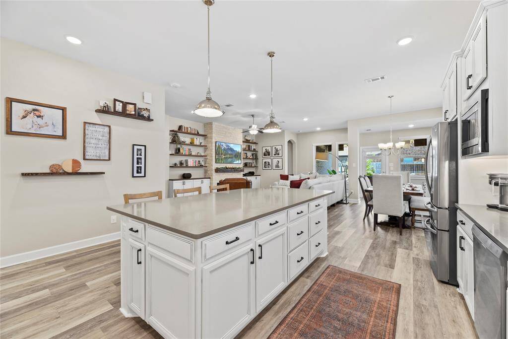 This spacious kitchen and living area features a large island with ample storage, modern pendant lighting, and stainless steel appliances. The open floor plan includes view lines of the living room with built-in shelves and a dining area with large windows for natural light. The light wood flooring and neutral walls create a bright and homey atmosphere.