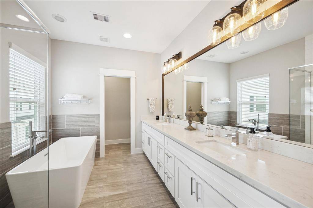 This gorgeous bathroom features a spacious dual-sink vanity with elegant lighting, a sleek freestanding bathtub, and a glass-enclosed shower. The neutral color palette and large windows create a bright, inviting atmosphere.