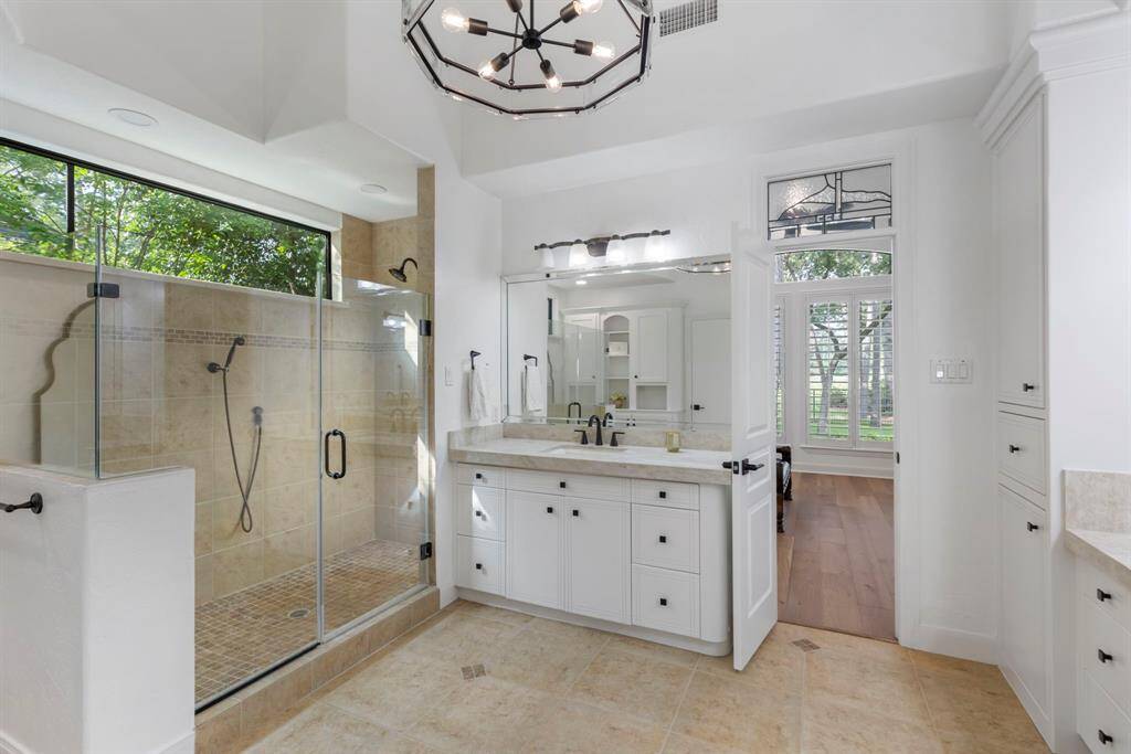 Elegant light fixtures and decorative framed mirrors enhance the bright, airy space, which also includes tiled flooring and an oversized walk-in shower with a glass enclosure and built-in accessory niche.