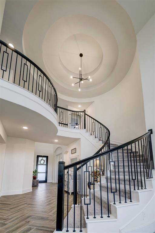 The grand foyer leads to magnificent staircase with elegant railing. The space is accented by a modern chandelier and herringbone-patterned flooring, creating a welcoming and luxurious entrance.