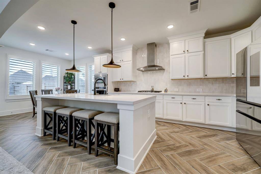 This modern kitchen features white cabinetry, a large central island with seating, and sleek stainless steel appliances. The space is bright with natural light from large windows and includes elegant pendant lighting. The floor has a stylish herringbone pattern, adding a touch of sophistication.