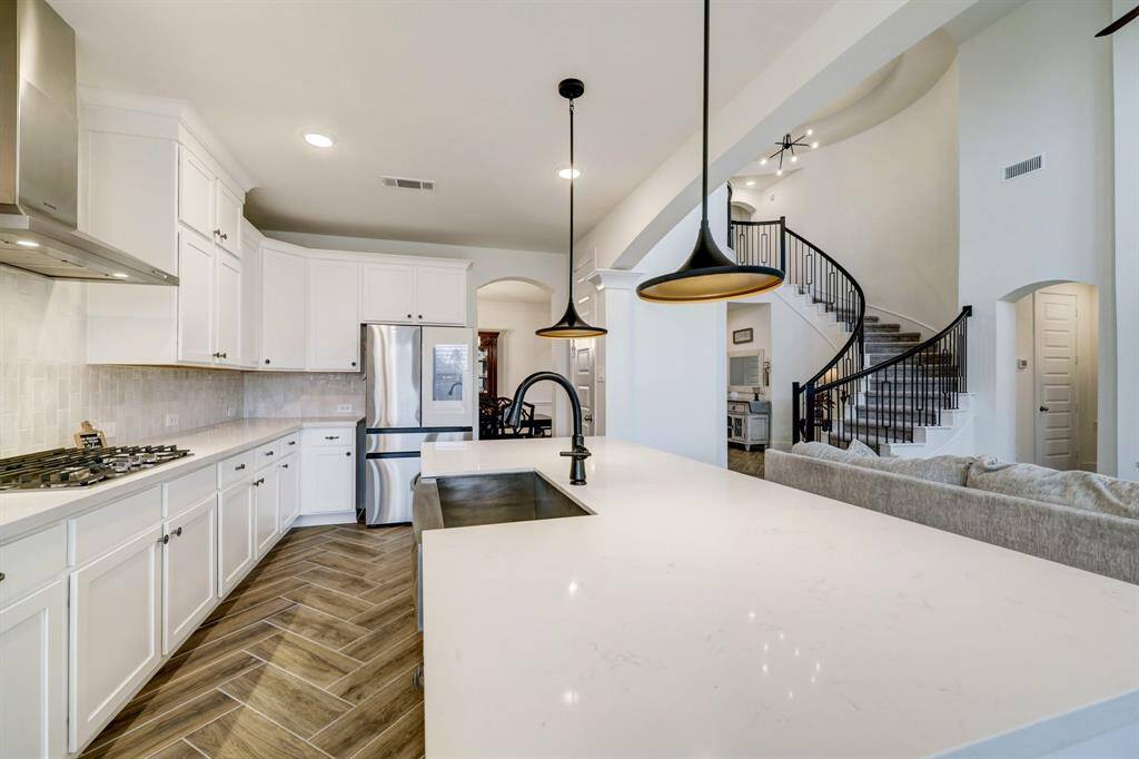This spacious kitchen features modern white cabinetry, stainless steel appliances, and a large island with elegant pendant lighting. It opens to a living area with a stylish spiral staircase and high ceilings, offering a bright and airy atmosphere.
