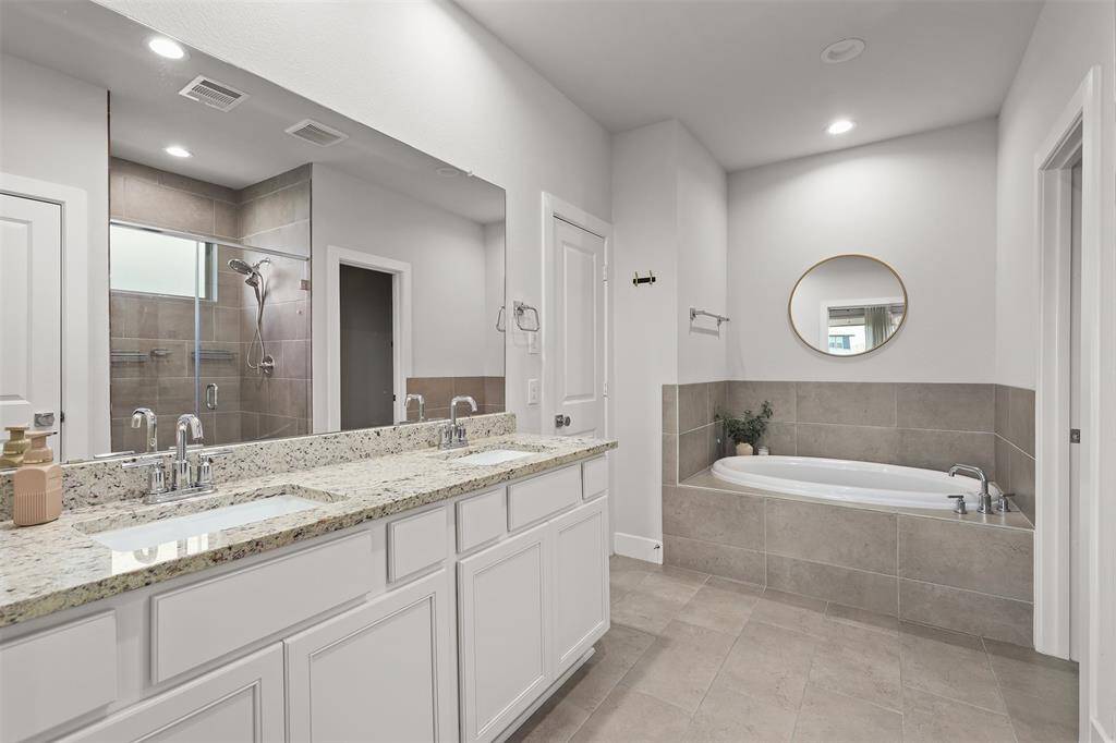 This modern bathroom features a spacious layout with a double-sink vanity topped with granite, a large mirror, and sleek fixtures. It includes a separate shower with glass doors and a soaking tub accented by neutral tile. Bright lighting and contemporary design create an inviting atmosphere.