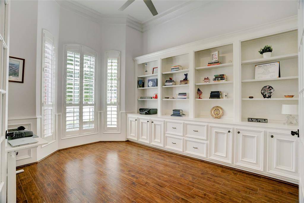 Bright and spacious home office with elegant built-in shelving, rich wood flooring, and large windows with plantation shutters, offering ample natural light and storage.