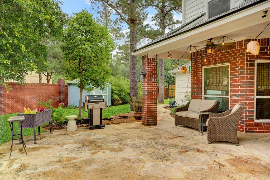Discover a charming backyard oasis featuring a spacious stone patio, perfect for cozy seating and outdoor dining with a grill. Surrounded by lush greenery and a privacy fence, it’s ideal for relaxation and entertaining under enchanting string lights.