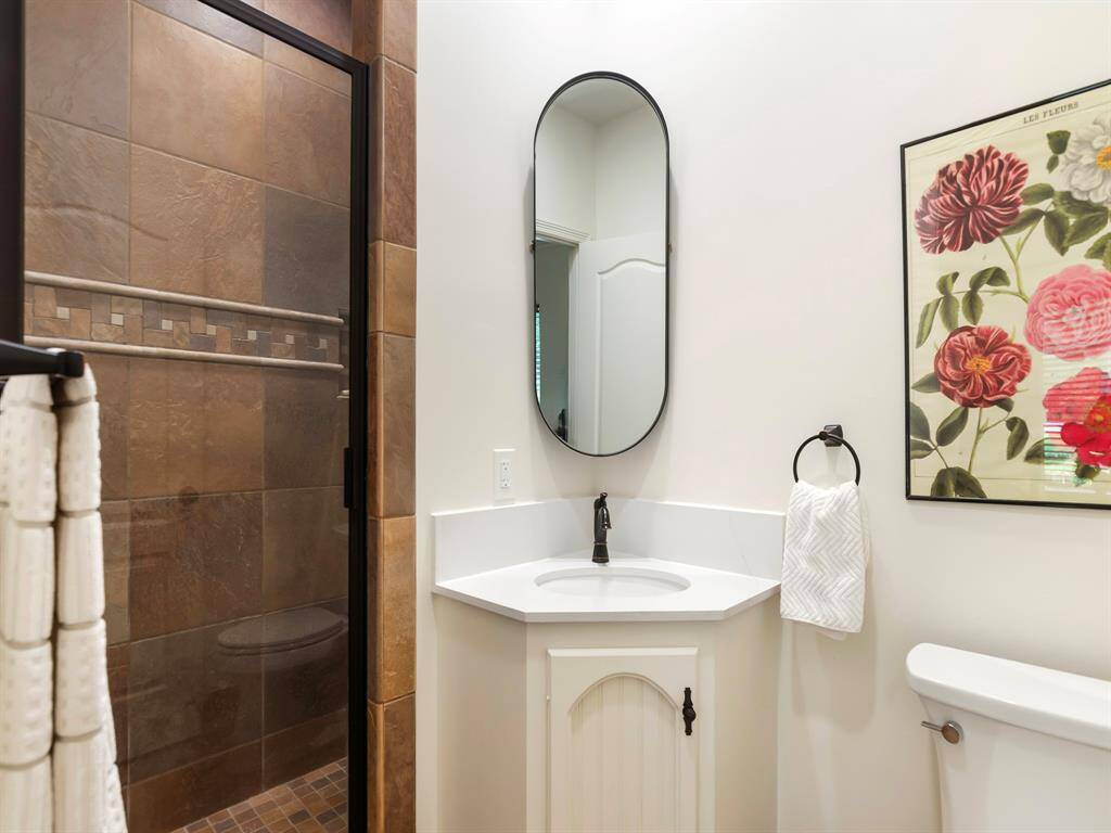 This bathroom features a stylish corner vanity with an oval mirror and a tiled shower with glass doors. The neutral tones and elegant fixtures create a welcoming and modern space.