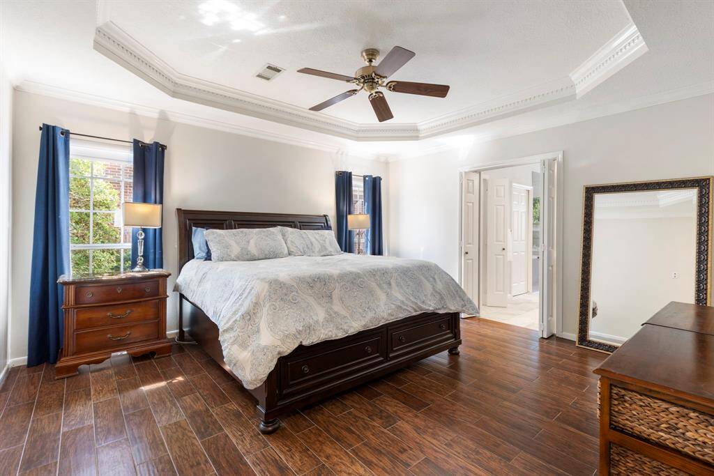 The primary suite is a serene retreat, featuring a tray ceiling with elegant dentil molding, a ceiling fan, and warm wood-look tile flooring that adds both comfort and style. A cleverly tucked-away accessory closet beneath the stairs (not pictured) includes Elfa shelving and a jewelry pull-out, offering smart and stylish organization.