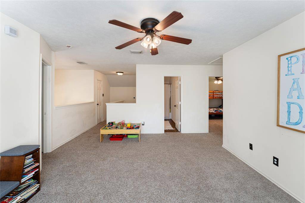 This spacious, carpeted room features a ceiling fan and ample natural light from a large window. It's currently set up as a play area with toys and has an adjacent room visible, ideal for a family space or game room.
