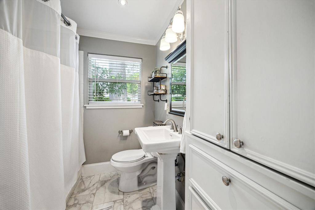 Bright window brings in natural light to this thoughtfully updated bathroom. Marble-look tile flooring complements the clean lines of the pedestal sink and black-framed mirror. Wall-mounted shelves offer both function and style, while chrome hardware and crisp finishes complete the look.