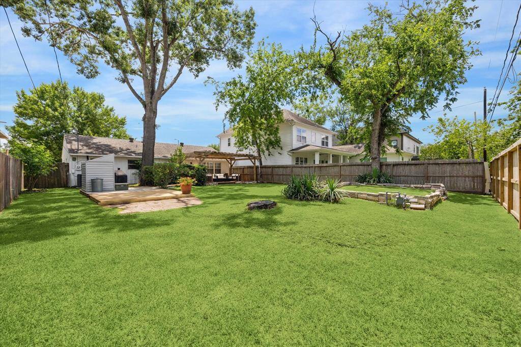 Wide-open yard with mature trees and layered landscaping offers endless possibilities for outdoor living. The raised platform and nearby fire pit create a natural hub for entertaining, while established garden beds and a pergola-covered patio round out this inviting green space.