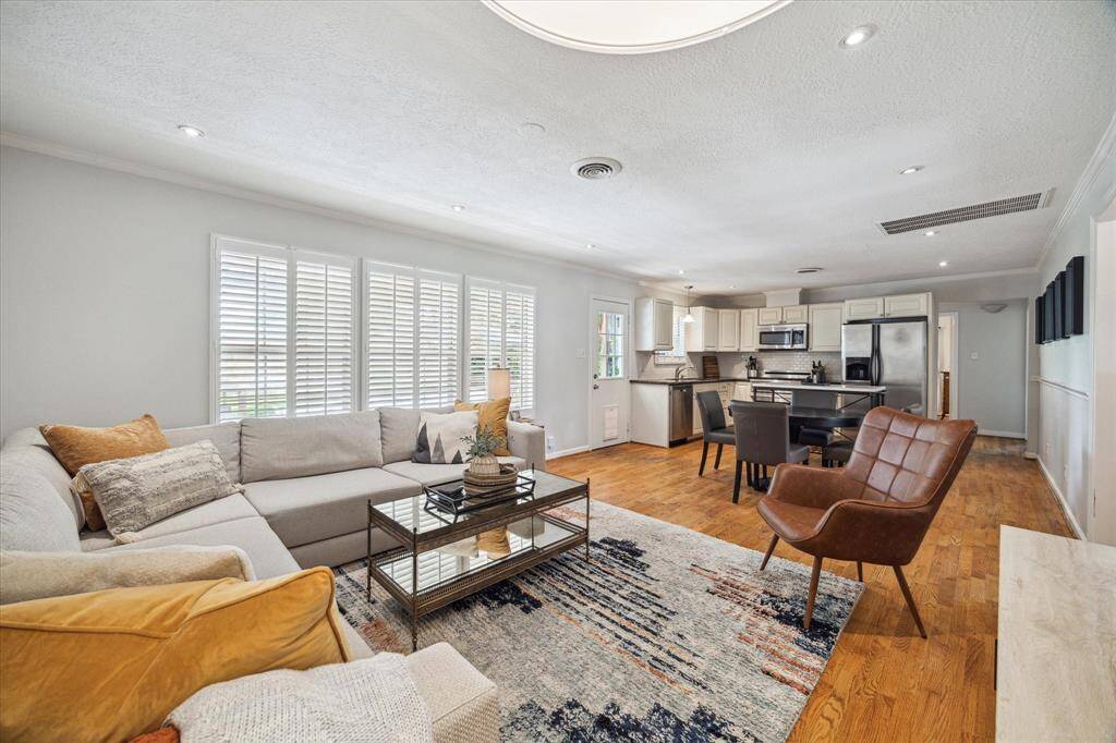Open-concept layout connects living, dining, and kitchen areas seamlessly. Natural light pours in through plantation shutters, highlighting wood flooring, neutral tones, and the flow between everyday living and entertaining spaces.