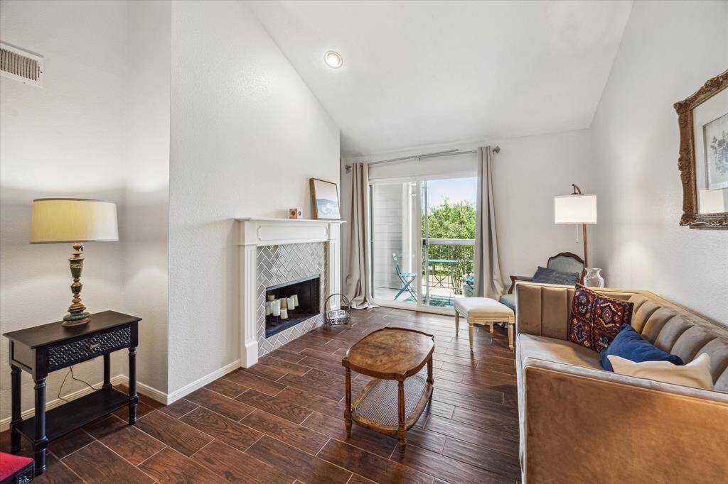 Soaring ceilings and fresh paint create an airy, welcoming feel in the spacious living room, where wood-look tile flooring adds warmth and durability. A custom-tiled fireplace with a handcrafted wood mantel serves as the focal point, flanked by a striking floor-to-ceiling built-in bookcase. Step through glass doors to a private balcony large enough to accommodate patio furniture for al fresco dining or relaxed evenings outdoors.