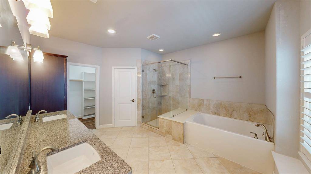 This spacious bathroom features a double vanity with granite countertops, a glass-enclosed shower, and a separate bathtub. A walk-in closet is accessible for added convenience.