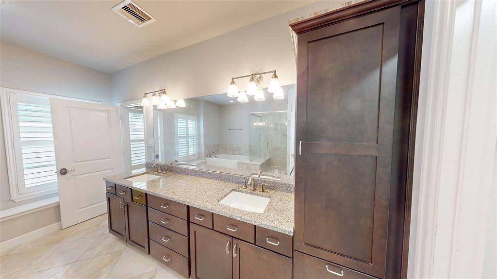 This dual-sink vanity features dark wood cabinetry for added storage. The room includes a large mirror, modern light fixtures, and a glass-enclosed shower. Natural light streams in through the window, enhancing the bright, airy feel.