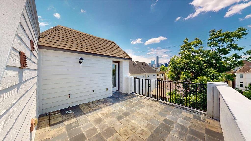 This photo showcases a spacious rooftop terrace with tiled flooring, offering a scenic view of the city skyline and surrounding greenery. The area is bordered by a black metal railing and has a charming exterior wall with a pitched roof and outdoor lighting. Perfect for relaxing or entertaining.