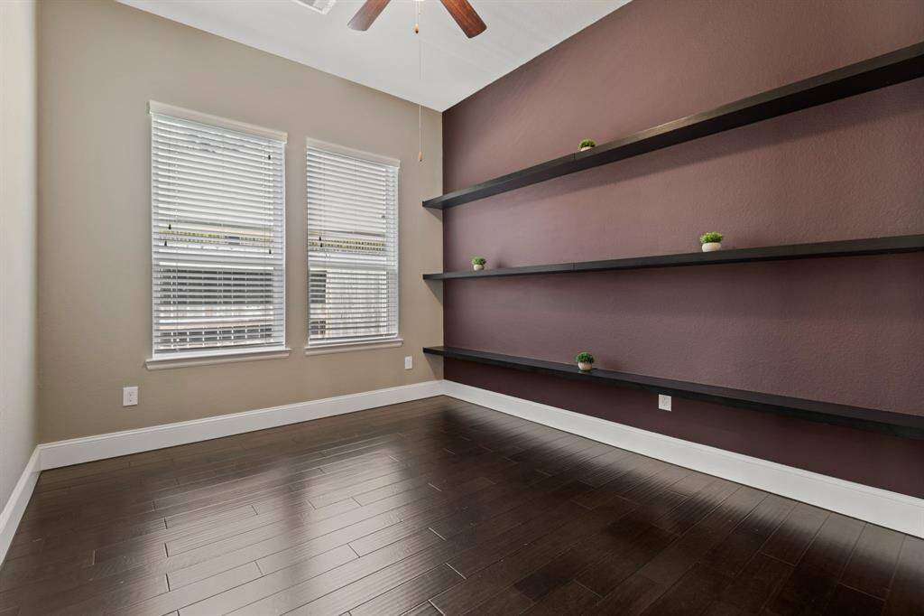 The Study/Flex Room features rich dark hardwood floors, a ceiling fan, and a bold accent wall with minimalist floating shelves. Two windows provide ample natural light, creating a bright and inviting space.