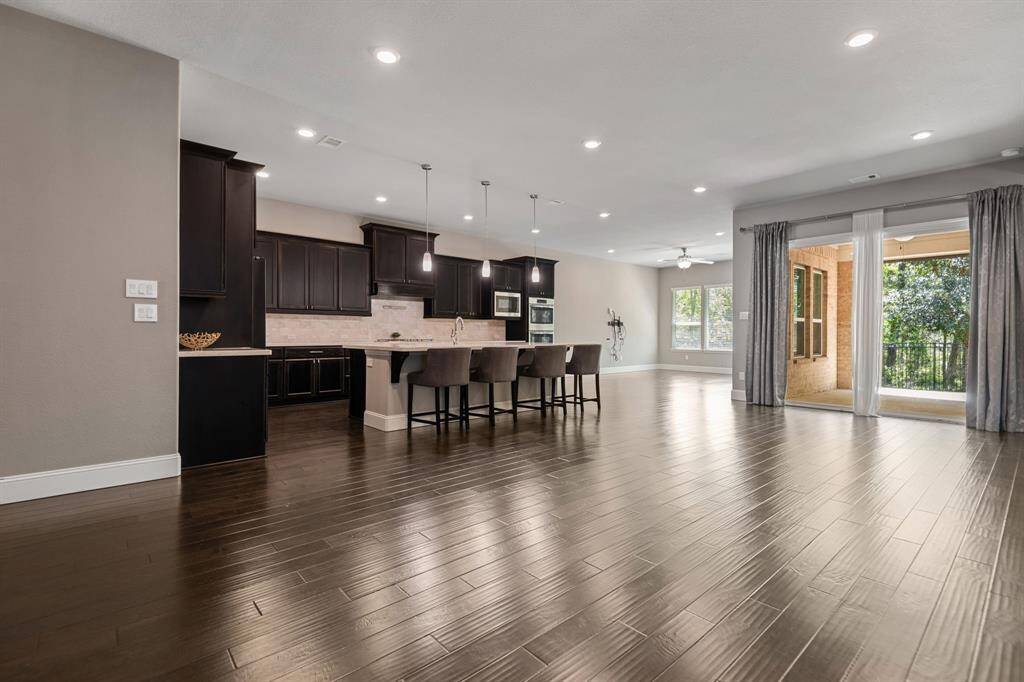 Spacious open-concept living area with modern kitchen, dark wood cabinetry, and large island with seating. Features hardwood floors, recessed lighting, and abundant natural light from large windows and sliding doors leading to a covered  patio. Perfect for entertaining.