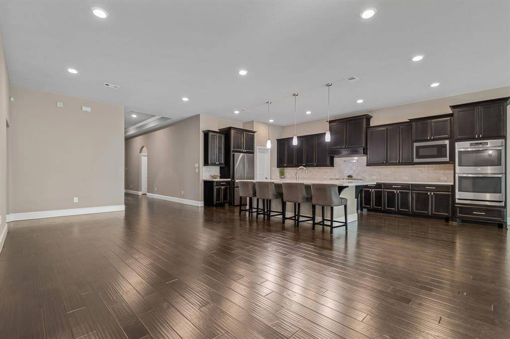 Spacious open-concept kitchen and living area featuring dark wood floors, modern dark cabinetry w/stainless steel hardware, stainless steel appliances, and a large island with seating. Recessed lighting and neutral walls create a bright, inviting atmosphere.