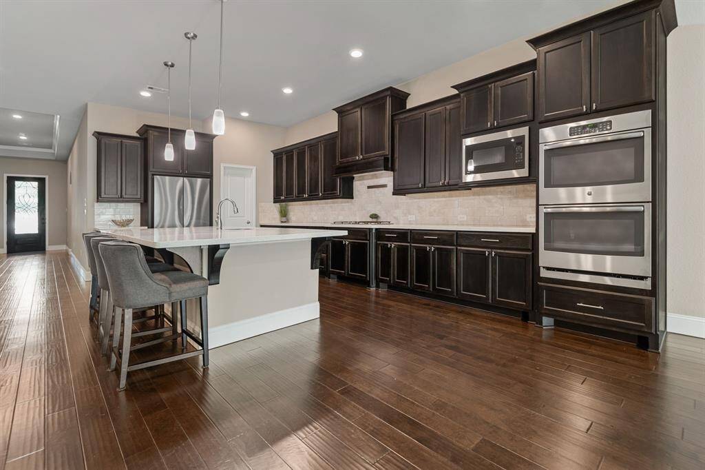 This spacious kitchen features dark wood cabinetry, stainless steel double ovens, microwave, cooktop and a large island with seating. The open layout, pendant lighting, and hardwood floors create a modern and inviting atmosphere.