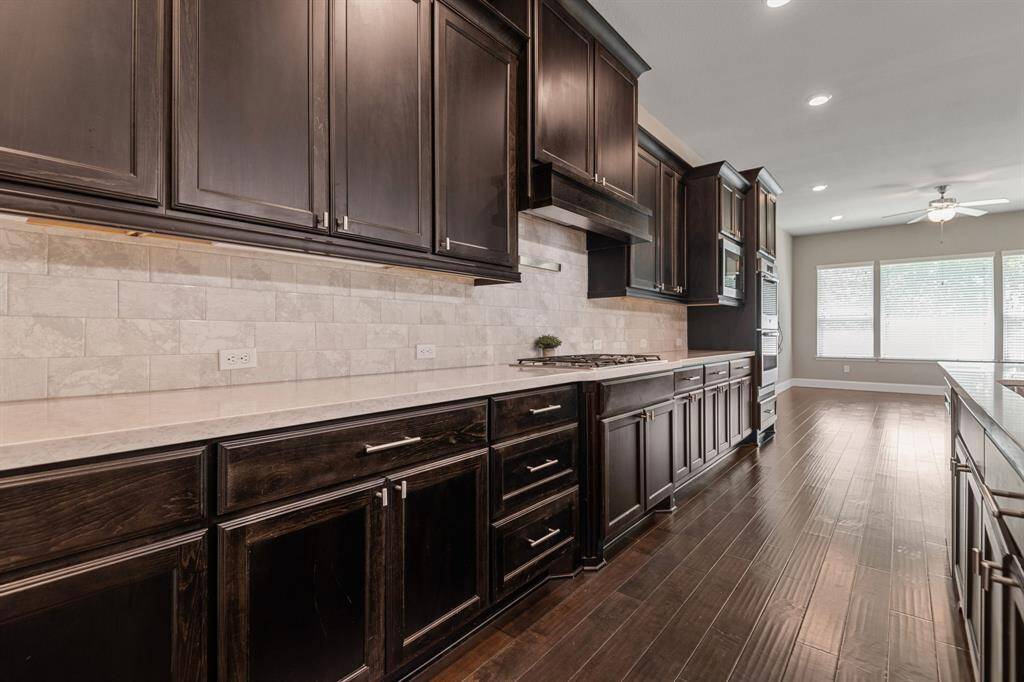 This kitchen features dark wood cabinetry, light countertops, and a tiled backsplash. It includes a built-in stovetop and modern appliances, with ample natural light from large windows and recessed ceiling lights. The space is open and inviting, ideal for cooking and entertaining.