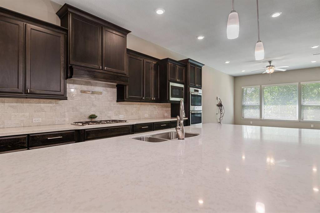 Modern kitchen with sleek dark cabinets and a spacious white countertop island. Features stainless steel appliances, an integrated cooktop, and stylish pendant lighting. Large windows provide ample natural light.