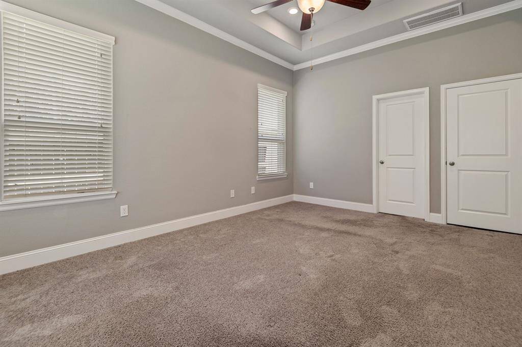 This photo shows another view of the Primary  Bedroom with neutral gray walls and carpet flooring. It features a ceiling fan, windows with blinds for natural light, and white double doors leading to the Primary Bath. The room has a clean, modern look.