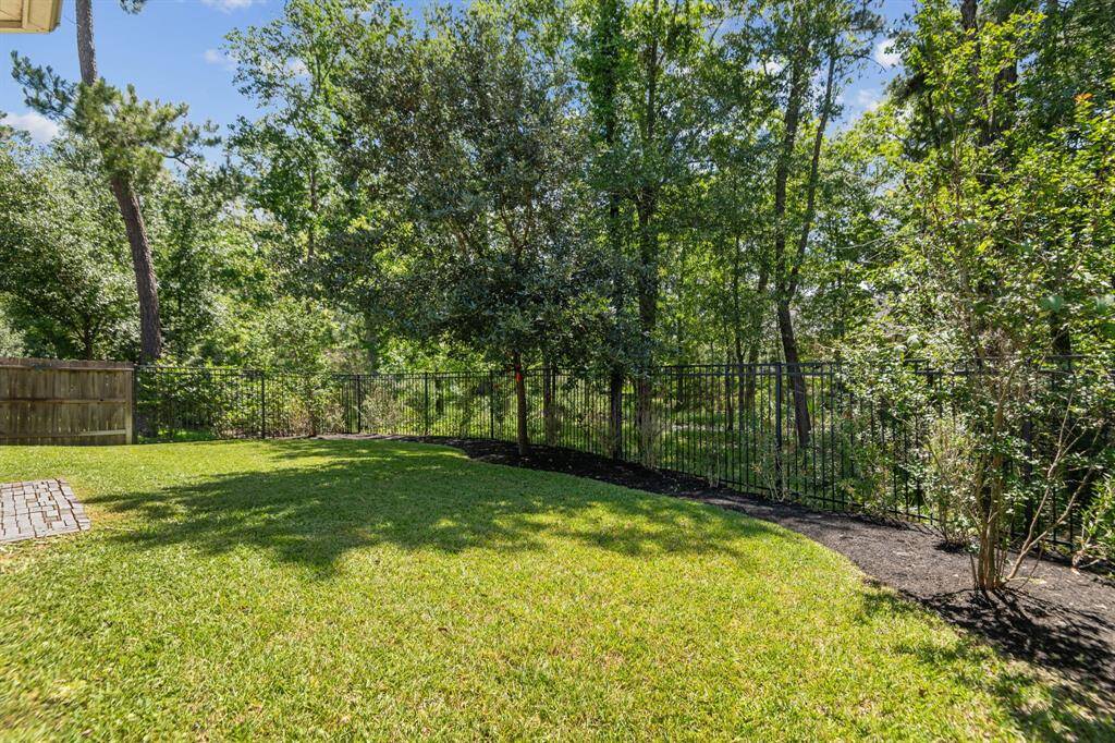 This photo showcases the spacious backyard with beautiful lawn, bordered by a wrought iron fence looking out onto the wooded greenbelt area offering privacy and a natural setting.