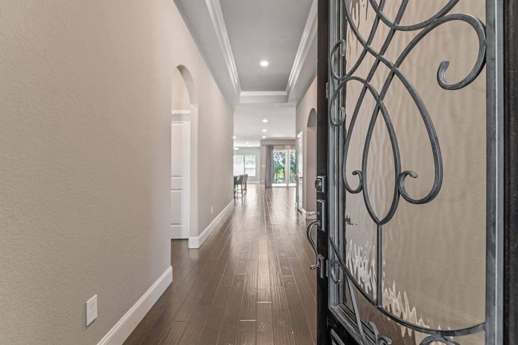 Welcoming entryway with elegant wrought iron door, leading to a bright hallway featuring rich hardwood floors and tray ceiling with recessed lighting .