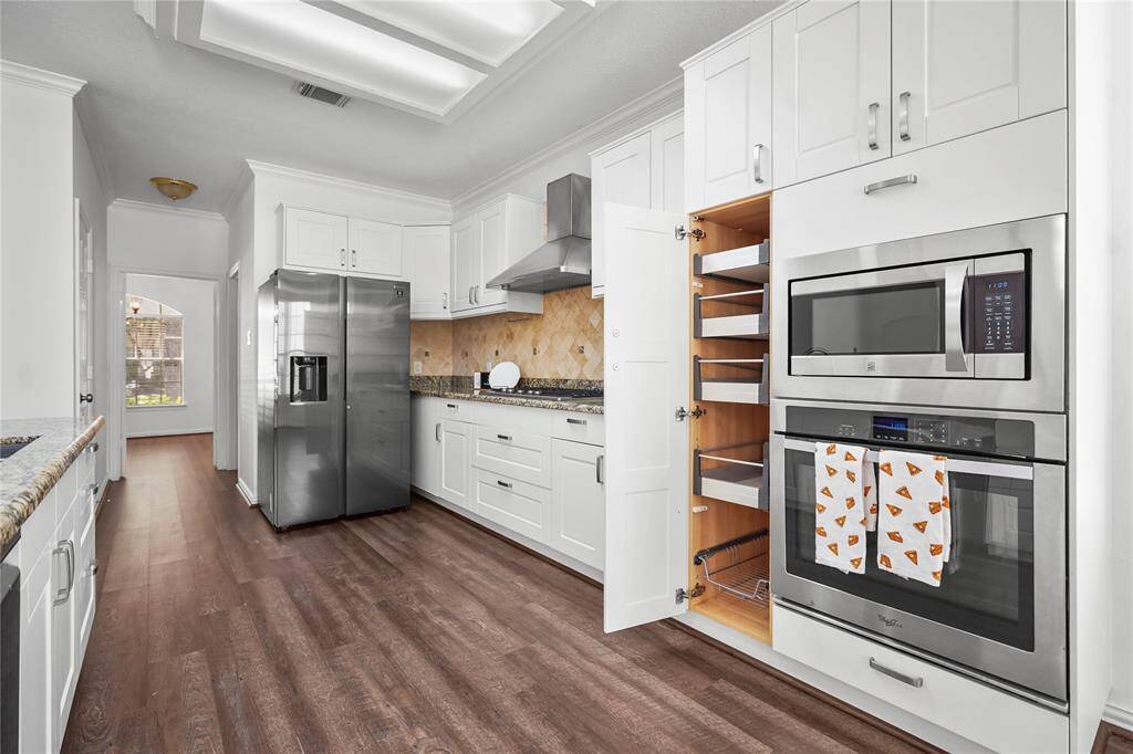 This modern kitchen features sleek stainless steel appliances, white cabinetry, and a pull-out pantry for convenient storage. The granite countertops and wood flooring add warmth and elegance, with ample natural light from the adjoining room.