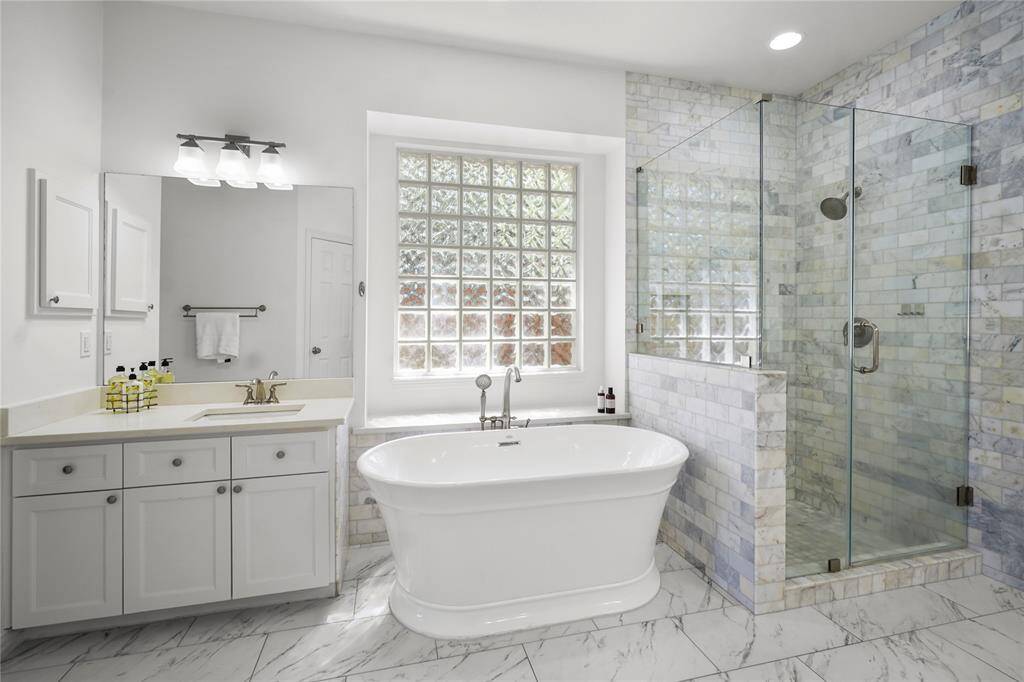 This bathroom features a luxurious freestanding tub, a spacious glass-enclosed shower, and elegant marble tile throughout. Ample natural light flows through the large glass block window, enhancing the clean and modern aesthetic.