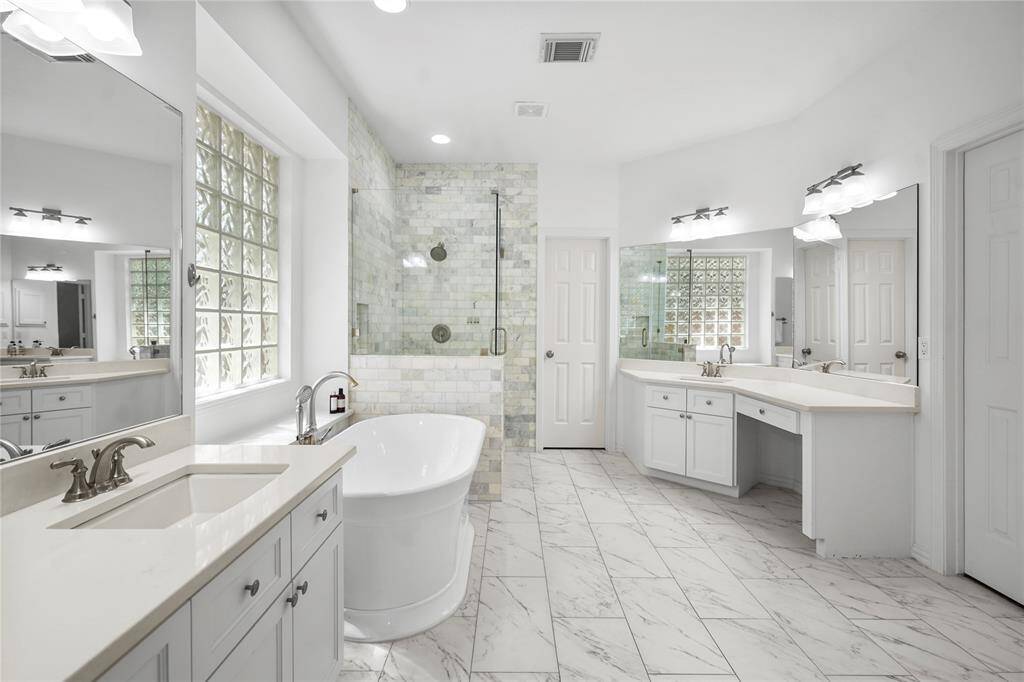 This photo showcases a spacious, elegant bathroom featuring a freestanding tub, a large walk-in shower with glass doors, dual sinks with ample counter space, and bright marble flooring. The room is well-lit with modern fixtures and natural light from a glass block window.