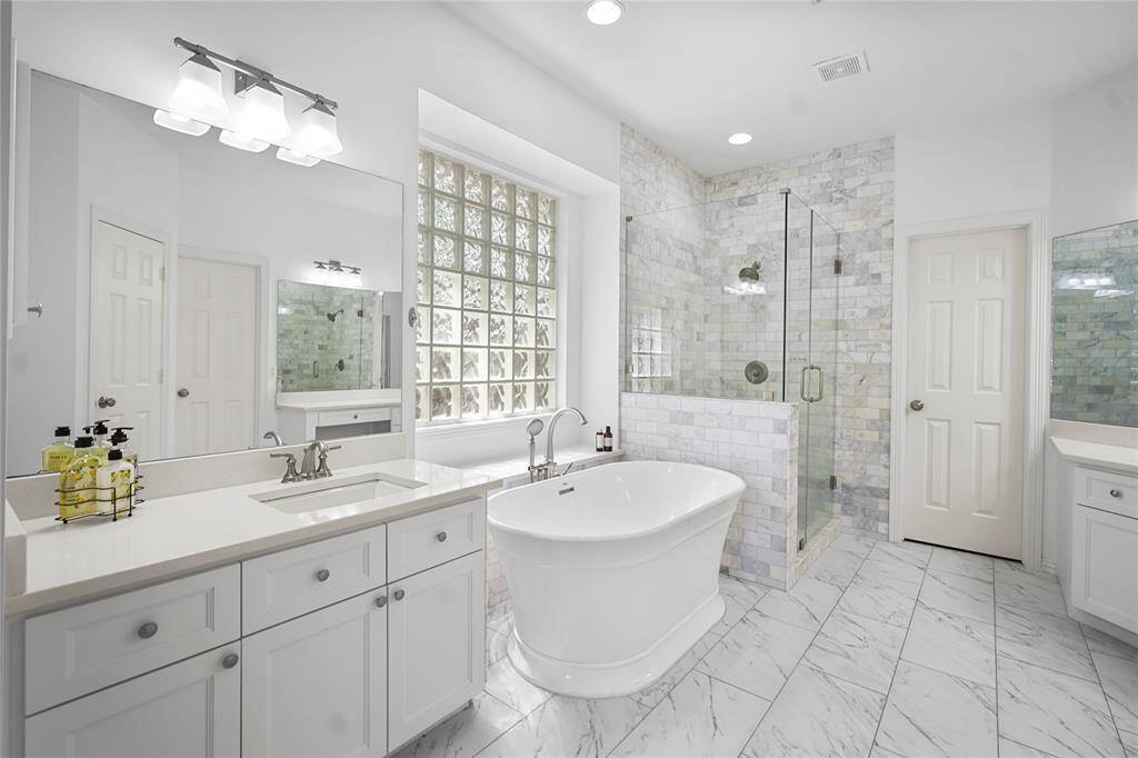This bathroom features a luxurious freestanding tub, a spacious glass-enclosed shower, and elegant marble tiles. The room is bright with ample natural light from a large glass block window, complemented by modern fixtures and white cabinetry for a clean, sophisticated look.
