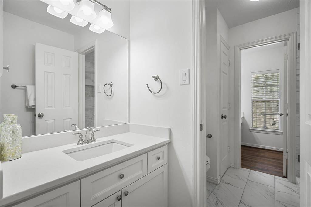 This bathroom features a modern design with a sleek white vanity, ample countertop space, and a large mirror. It has elegant lighting, marble-style flooring, and a window that provides natural light.