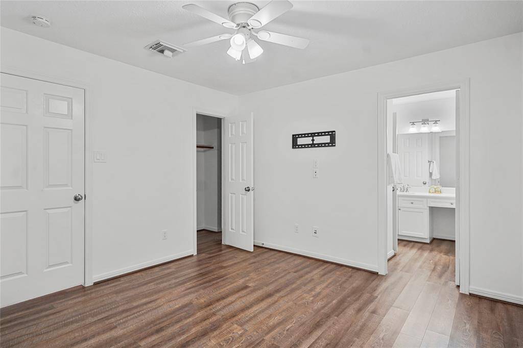 This photo shows a bright, white-walled room with wood flooring, a ceiling fan, and a mounted TV bracket. It features a walk-in closet and an adjoining bathroom with a vanity and ample lighting.