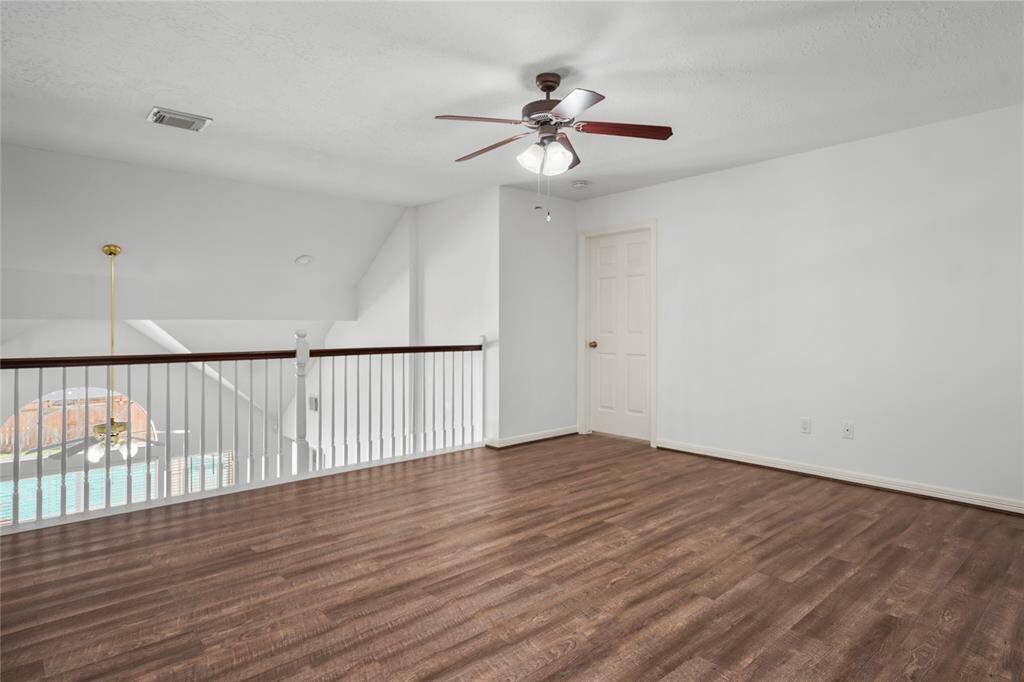 This photo showcases a bright, open loft space with hardwood floors, a ceiling fan, and a view of an indoor pool area. The white walls and railing add to the airy feel, making it a versatile area for a home office or lounge.