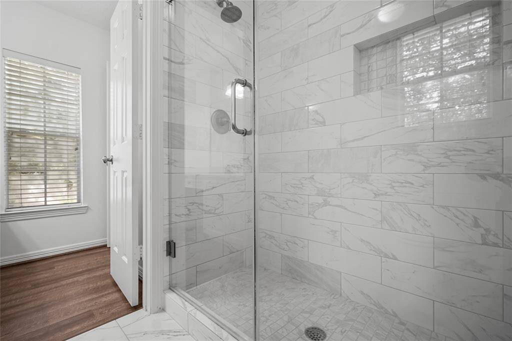 This photo showcases a modern bathroom with a glass-enclosed shower featuring elegant marble-like tiles. Natural light filters through a window with blinds, complementing the sleek, clean design.