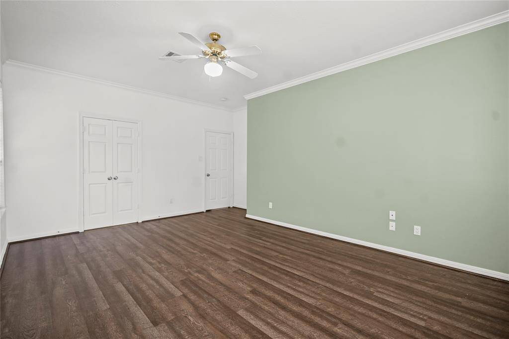 This spacious room features dark wood flooring, a light green accent wall, and a white ceiling with a ceiling fan. It includes two white doors and ample natural light, offering a versatile living space.