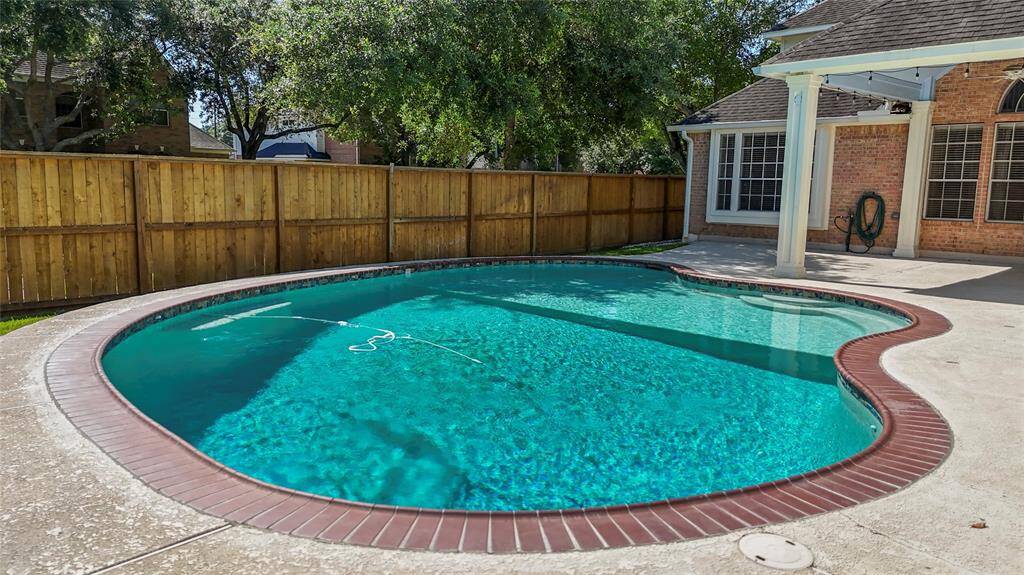 This photo showcases a charming backyard with an inviting swimming pool featuring a red brick border. It's surrounded by a wooden privacy fence and shaded by mature trees, offering a serene outdoor space. The adjacent patio area is perfect for relaxing or entertaining.