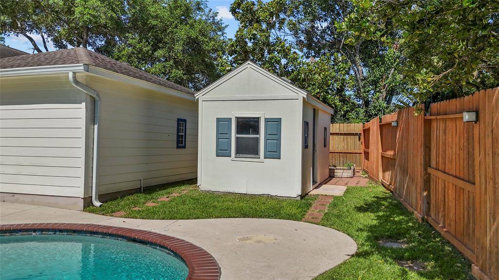 This photo features a backyard with a small shed adjacent to a larger building, bordered by a wooden fence. The area includes a grassy lawn and a paved walkway leading to the shed. A pool with a brick edge is partially visible, adding to the outdoor amenities.