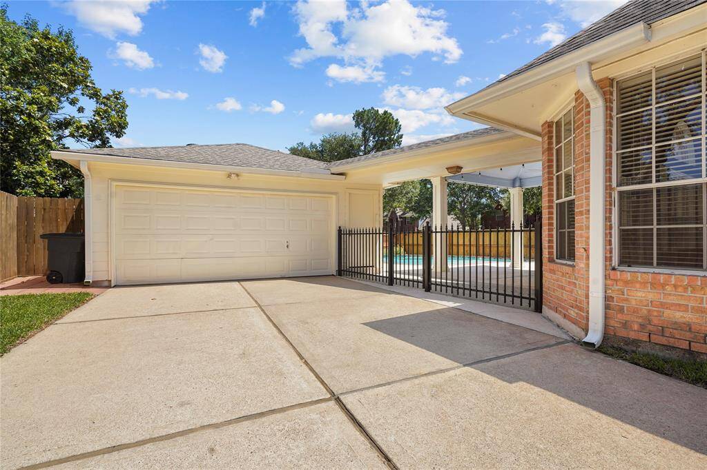 This photo showcases a spacious driveway leading to a two-car garage, adjacent to a brick house with large windows. There is a gated entrance to a covered patio area, offering a view of a pool in the backyard, surrounded by trees and fencing for privacy.