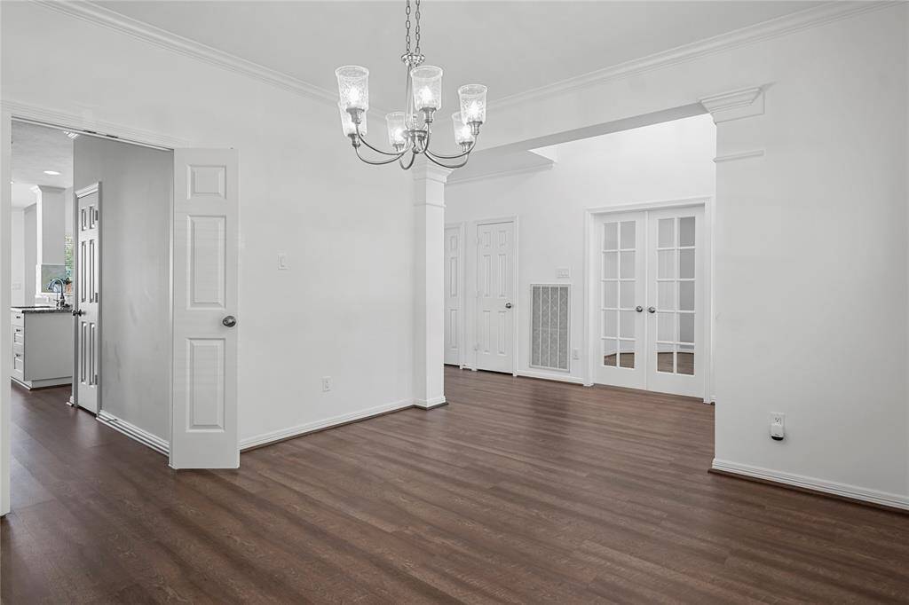 This room features elegant hardwood flooring and bright white walls, creating a spacious and inviting atmosphere. The chandelier adds a touch of sophistication, while French doors and crown molding enhance the classic appeal. A glimpse into the adjacent kitchen area suggests a seamless flow between spaces.