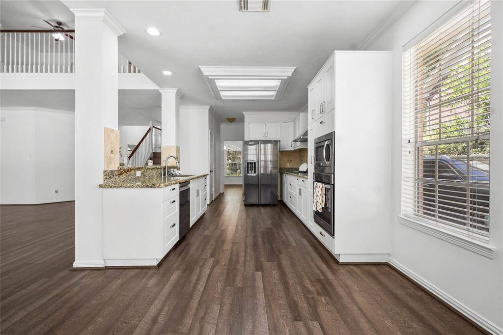 This modern kitchen features sleek white cabinetry, granite countertops, and stainless steel appliances. It has ample natural light from large windows and a spacious layout with hardwood flooring, offering a clean and inviting atmosphere.