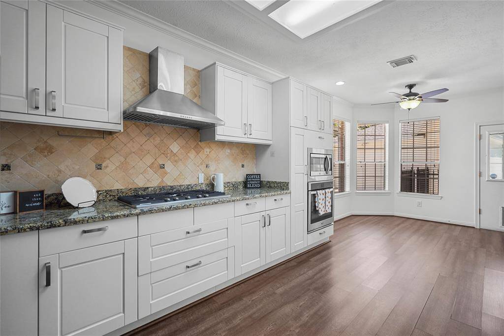 This kitchen features modern white cabinetry, a stylish backsplash, and granite countertops. It includes a built-in oven, microwave, and a sleek range hood. The space is bright with large windows and wood flooring, offering a cozy dining area with a ceiling fan.