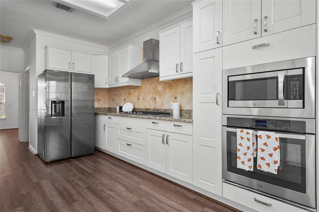 Modern kitchen with stainless steel appliances, white cabinetry, and granite countertops. The room features a built-in oven, microwave, and a spacious refrigerator. The wood flooring adds warmth to the sleek design.